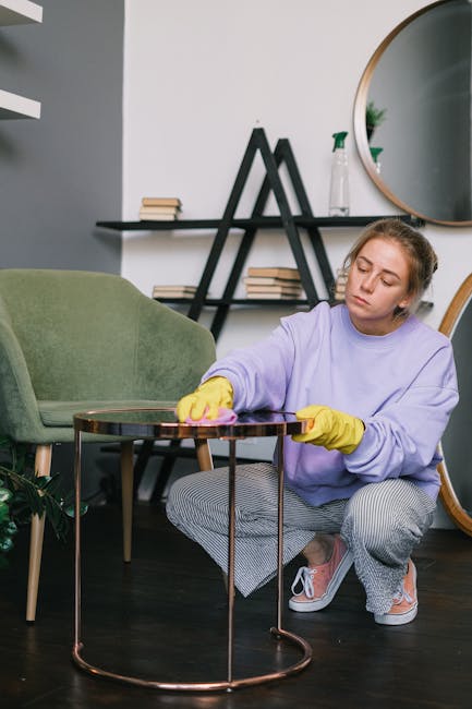 A young woman with light skin, wearing a lavender long-sleeve top, yellow cleaning gloves, and grey and white striped pants, is kneeling on a dark wooden floor in a modern living room. She is polishing a small round brown side table with a pink cloth, demonstrating surface cleaning and maintenance. The room features a green upholstered armchair with wooden legs, a black triangular shelving unit against the white wall holding books and decorative items, and a large circular mirror. Bright lighting highlights the clean, dust-free surfaces, emphasizing hygiene and deep cleaning practices. The setting reflects a professional domestic cleaning service provided by Carpet Cleaners Harringay, ensuring clean and maintained living spaces.