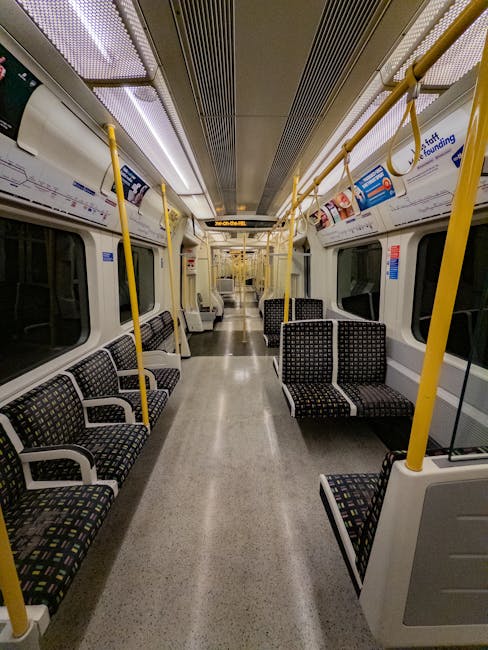 An empty train carriage interior with rows of black upholstered seats featuring multicolored geometric patterns, positioned along both sides of the aisle. The carriage has a clean, polished light grey floor and white walls with large windows, framed by black trim. Bright ceiling panels with integrated lighting and stainless steel handrails with yellow vertical supports run parallel to the seating. Advertisements and informational posters are visible above the windows. The overall scene emphasizes a maintained, hygienic environment, reflecting professional cleaning practices. Carpet Cleaners Harringay specializes in surface cleaning and deep cleaning for domestic and commercial spaces, ensuring high standards of sanitisation.