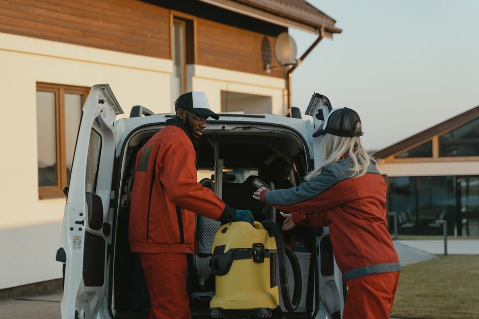 A female cleaning technician wearing a red and gray uniform stands in front of a white service van parked on a paved driveway outside a modern wooden house. She holds a vacuum cleaner in her right han