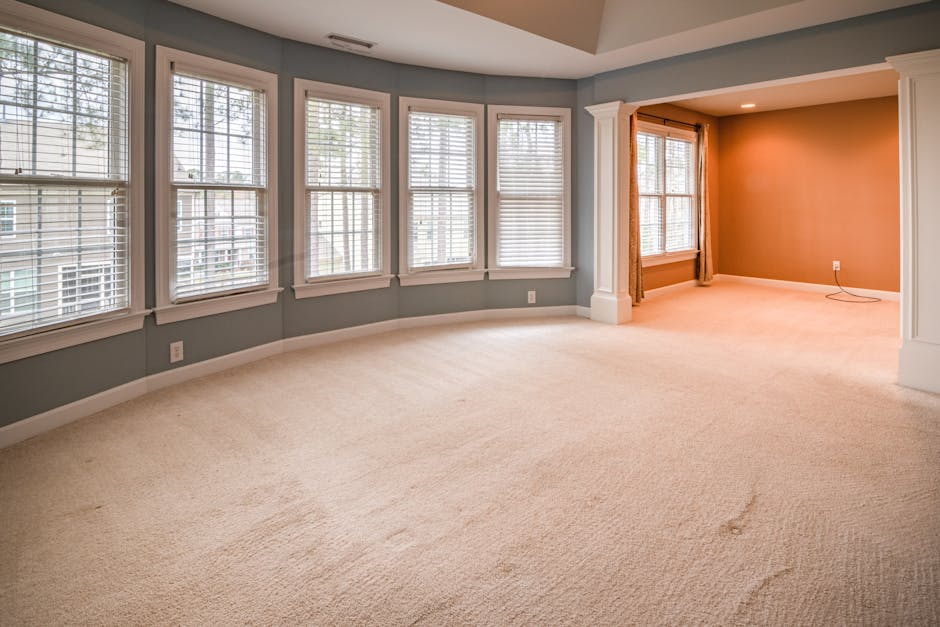 A modern living room with large floor-to-ceiling windows showcasing a cityscape view, featuring white walls and a light hardwood floor. The room contains a spacious, light gray sectional sofa with a s