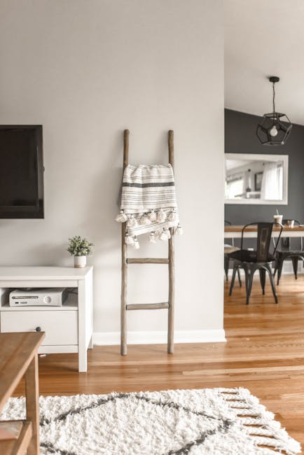 The image depicts a modern living space with a light-colored wall and hardwood flooring. There is a wooden ladder-style coat rack positioned centrally against the wall, with a striped cloth towel hanging on one of its rungs. To the left of the ladder, a small white console table holds a potted plant, and a flat-screen TV is mounted on the wall above it. The foreground features a textured, plush white rug on the wooden floor. In the background, part of a dining area with a wooden table and black chairs is visible, illuminated by natural light from windows and complemented by a ceiling pendant light. The room appears clean and well-maintained, reflecting professional surface cleaning and domestic cleaning standards that Carpet Cleaners Harringay offers for homes in Harringay Ladder, ensuring hygiene and surface freshness.