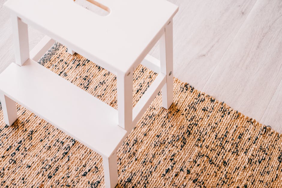 Close-up view of a white wooden step stool placed on a woven beige and black speckled rug in a room with light wooden flooring. The surface of the stool appears clean and well-maintained, reflecting attention to hygiene and surface cleaning. Damp or polished surfaces are not visible, and no dust or debris is present. The lighting is natural and soft, highlighting the tidy condition of the area, which is part of a domestic setting where Carpet Cleaners Harringay provides deep cleaning and sanitisation services for rugs and upholstery to ensure optimal cleanliness and hygiene.