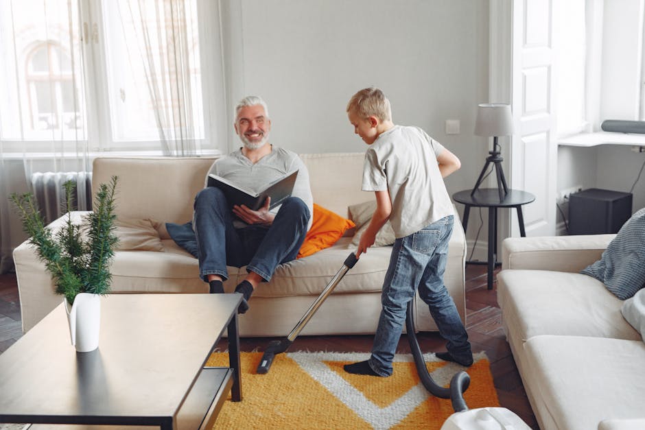 A young boy using a vacuum cleaner to clean an orange and white patterned rug in a bright living room, where an older man with grey hair and a beard sits on a beige sofa reading a book. The room features a white wall with large windows allowing natural light, a small black side table with a white lamp, a white armchair with a grey cushion, and a white side table with a potted evergreen plant. Carpet Cleaners Harringay is providing surface cleaning and hygienic maintenance in this residential space, highlighting the importance of regular sanitisation and deep cleaning to maintain cleanliness and hygiene in the home.