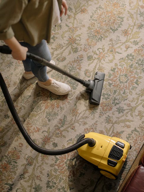 A child is standing on a patterned area rug with intricate floral designs in muted tones of beige, green, and orange, while using a yellow canister vacuum cleaner with a black hose attachment to clean the surface. The child's legs are visible, wearing blue jeans and beige sneakers, and they hold the vacuum's handle, guiding it across the rug. The room features soft, natural lighting, highlighting the clean, well-maintained appearance of the carpet. The image reflects professional deep cleaning and surface sanitation, with Carpet Cleaners Harringay's expertise implied through the cleaning activity on a residential rug, demonstrating the process of domestic surface cleaning with a focus on hygiene and maintenance.