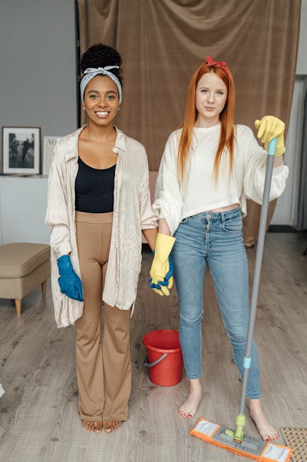 Two women standing indoors in a well-lit room, preparing for cleaning. The woman on the left has dark skin, curly hair tied with a light blue headband, and is dressed in a beige oversized shirt, black top, and tan trousers, wearing blue cleaning gloves. The woman on the right has fair skin, long red hair with a pink headband, wearing a white sweater and blue jeans, holding a mop with a yellow glove on one hand and standing barefoot. Behind them, a red bucket and cleaning tools are visible on the wooden floor. The room features a beige curtain, framed photos on the wall, and a grey upholstered chair, illustrating an environment ready for comprehensive surface cleaning, deep cleaning, and sanitisation, as provided by Carpet Cleaners Harringay.
